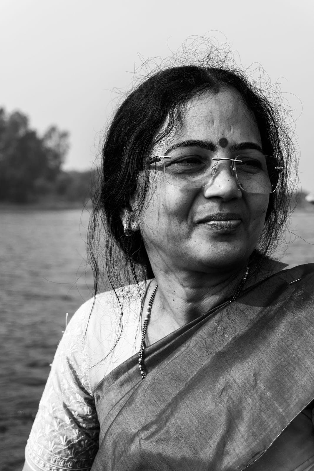 Black-and-white portrait of a smiling woman in glasses and a sari, outdoors by water
