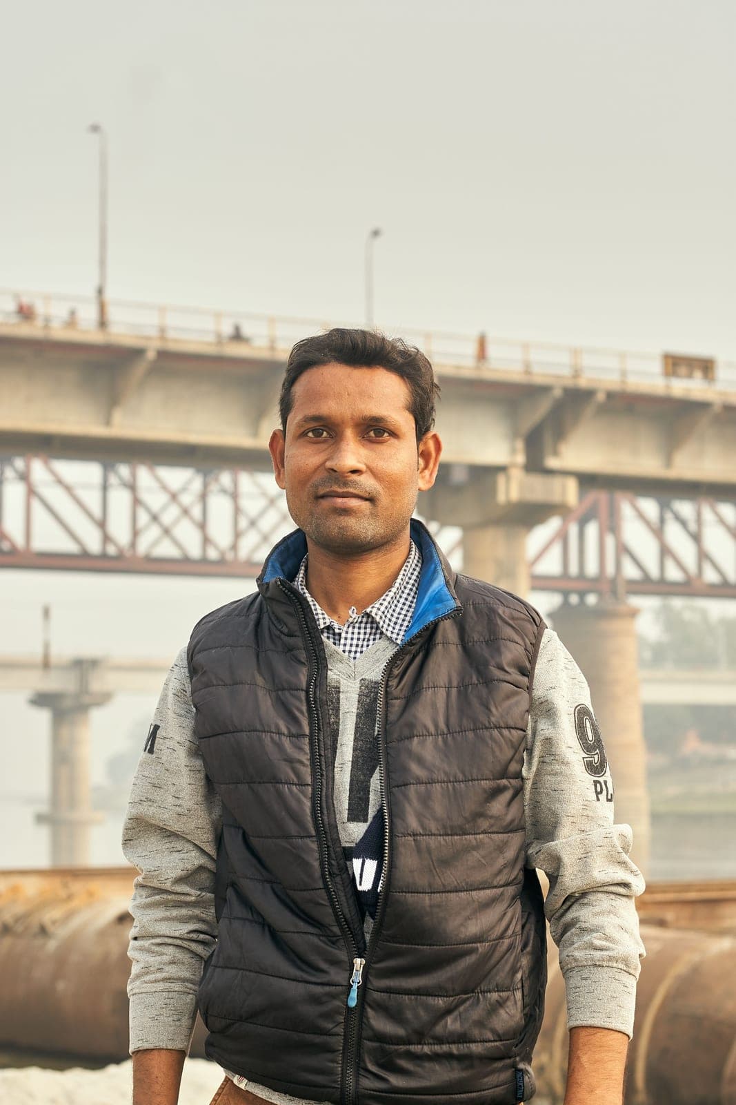 A man in a black quilted vest stands in front of a steel bridge on a hazy day