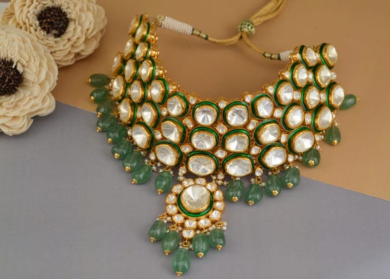 A kundan necklace with green bead drops, laid on a sandy ground beside crochet flowers