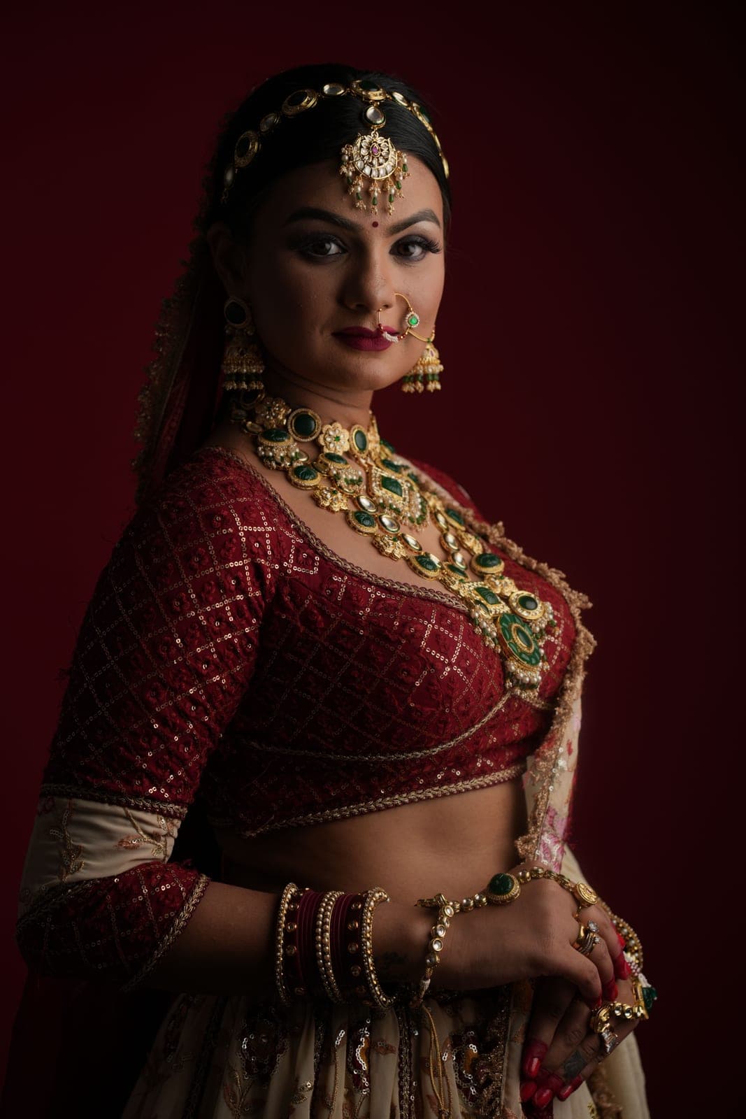 A bride in a maroon lehenga with a kundan rani haar and matha patti, against a red wall