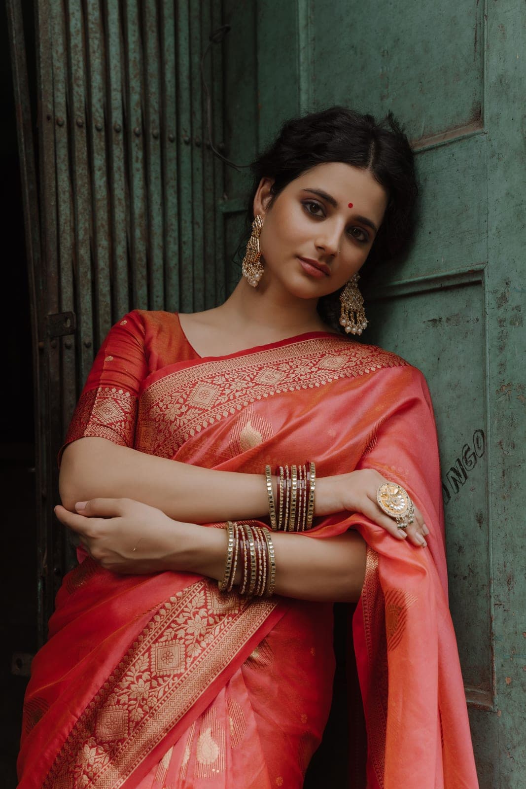 The model in the coral Banarasi leaning in a doorway painted green, bangles stacked to the elbow