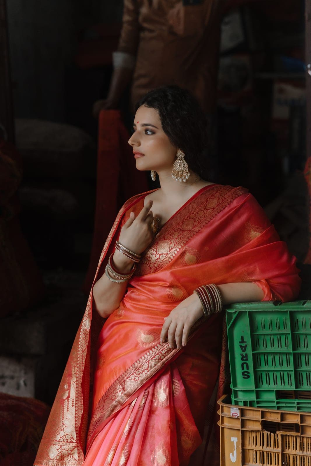 A model in a coral Banarasi saree with gold buti and glass bangles, beside market crates