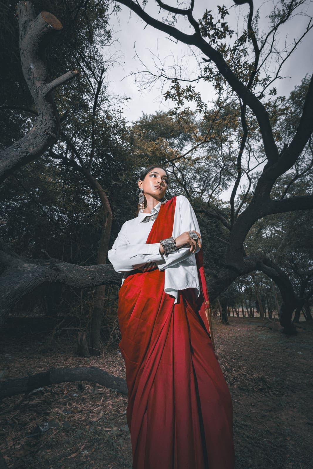A model in a red silk saree over a white cotton shirt, standing under old trees