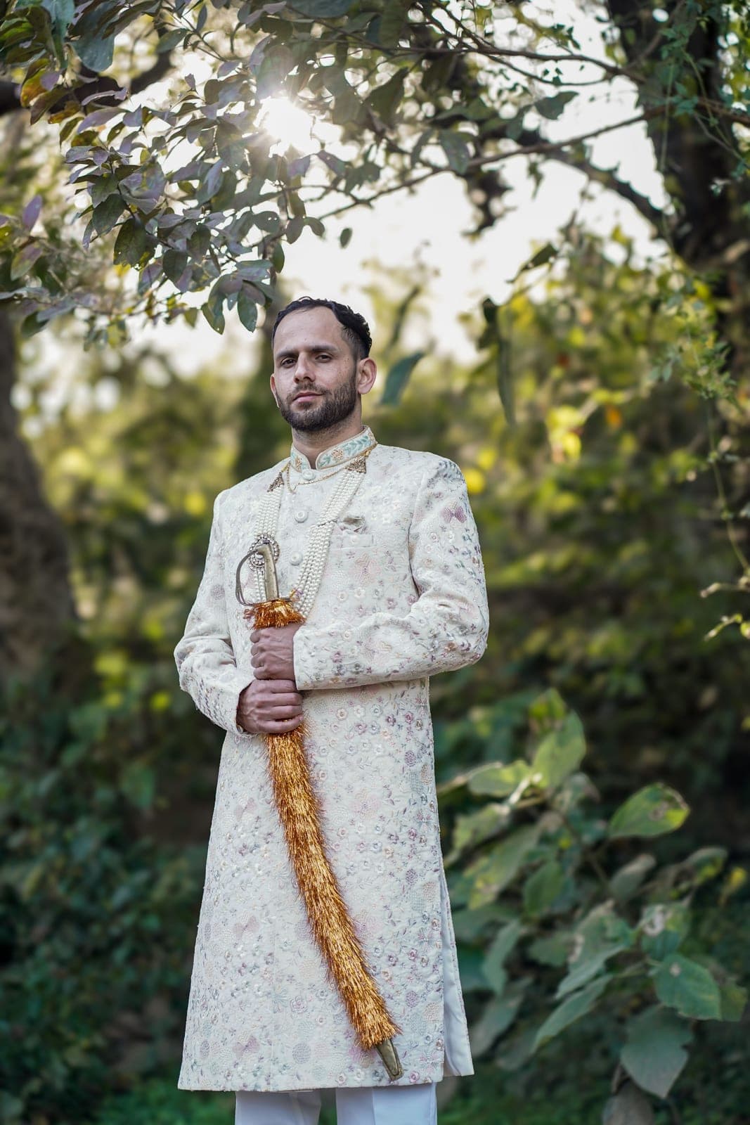 A groom in an ivory sherwani holding a sword with a marigold tassel, among trees