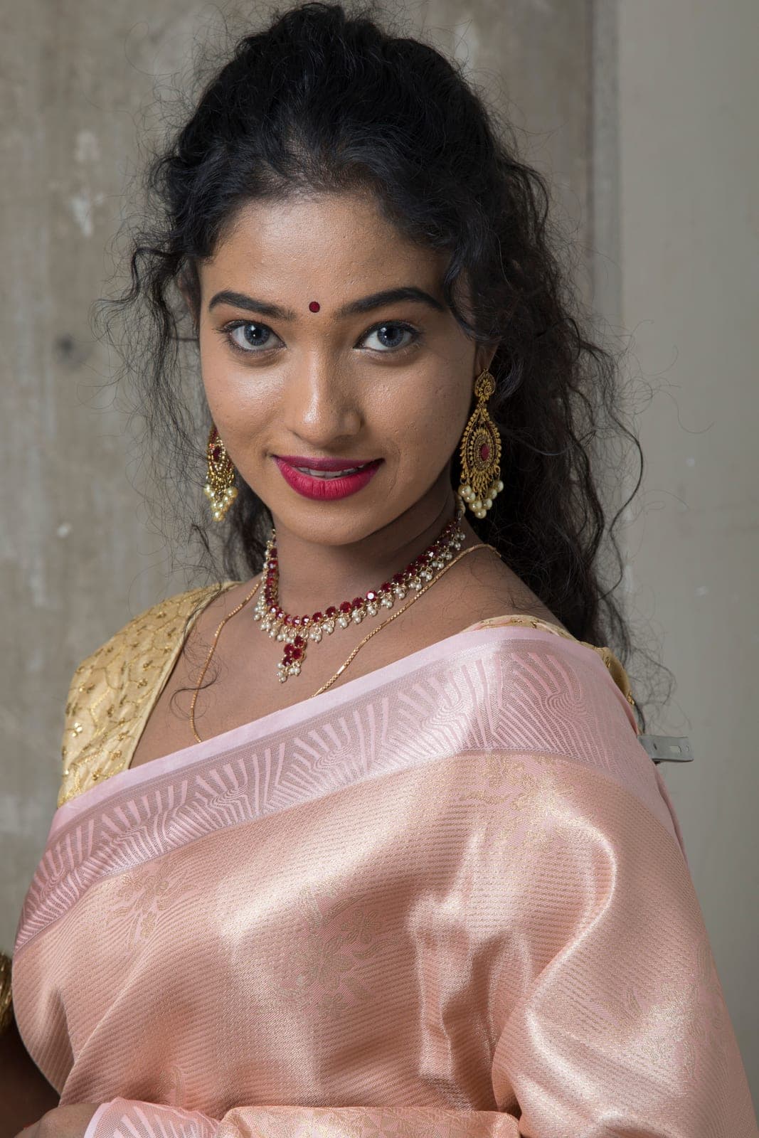 A woman in a rose-pink tissue saree with a ruby and pearl necklace and gold drop earrings