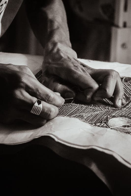 Black-and-white close-up of hands at an embroidery frame, holding the needle like a pen