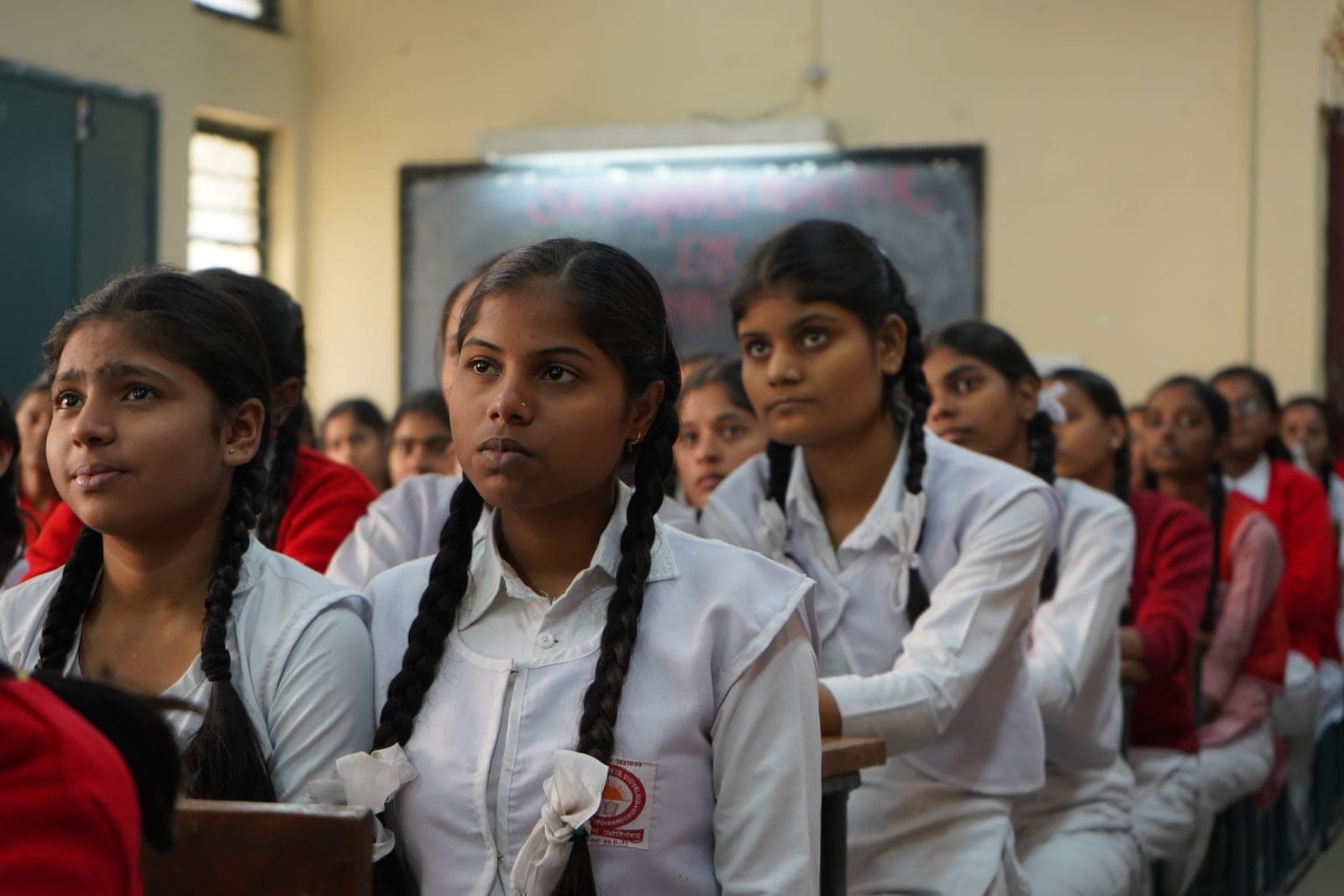 Girls in white uniforms at desks in a classroom, one standing to answer