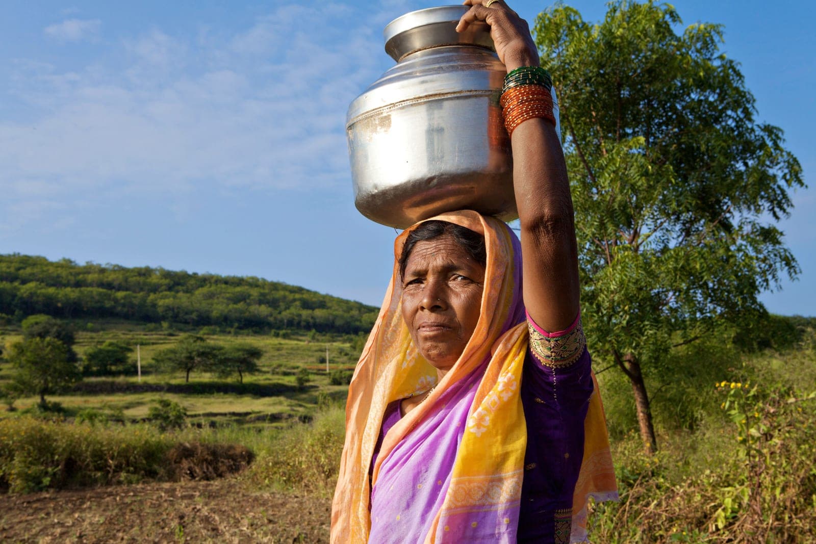 A woman in a sari carrying a brass water pot on her head along a village lane