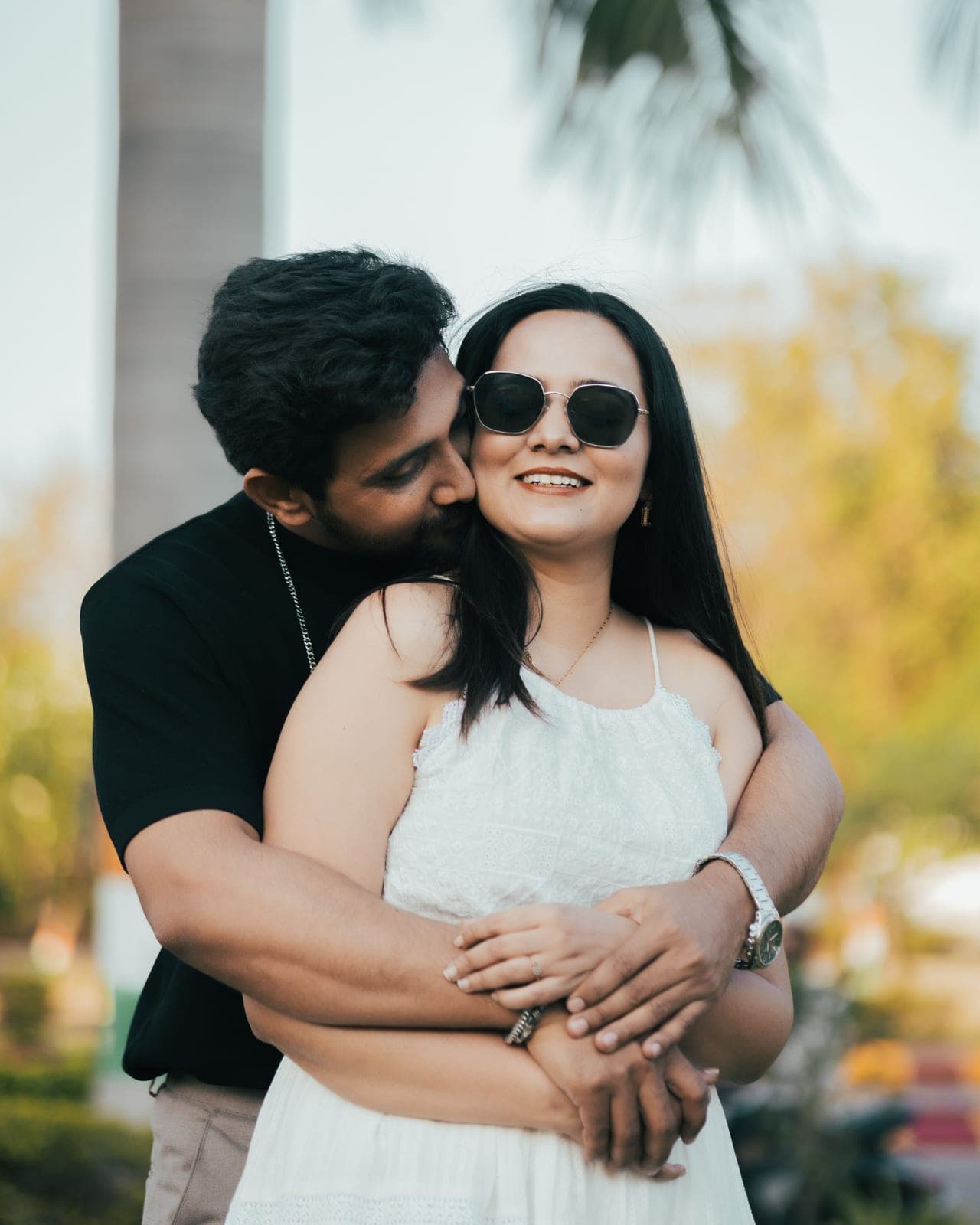 Arjun kisses Tanvi on the cheek; she smiles in sunglasses, trees soft behind them