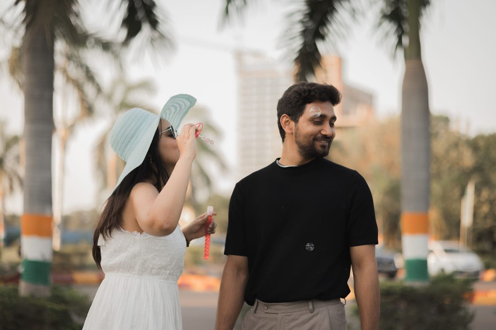 Tanvi blows soap bubbles under the palms while Arjun, in a black T-shirt, watches and smiles