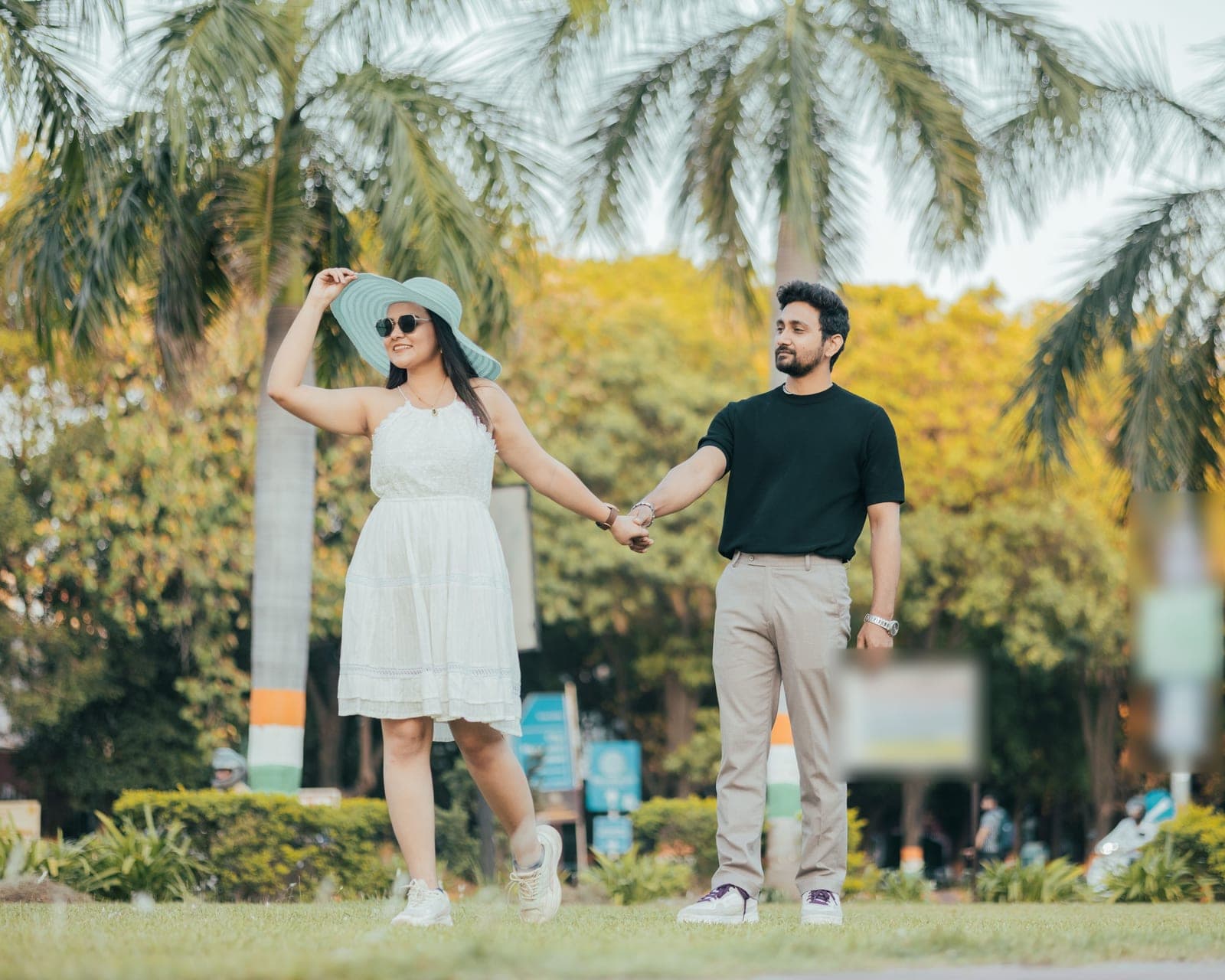 Tanvi in a wide sunhat and a white dress walks hand in hand with Arjun in a black T-shirt along a park path